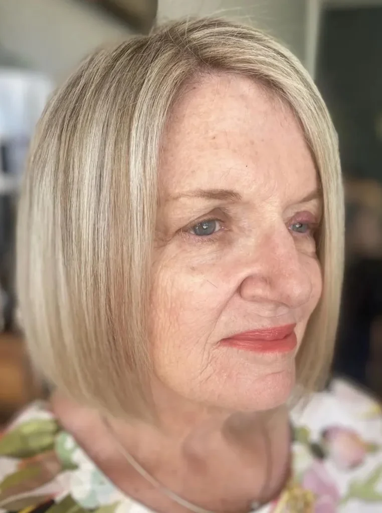 Frau über 60 mit blondem Bob und tiefem Seitenscheitel, glatte kinnlange Haare für einen eleganten, modernen und verjüngenden Look.