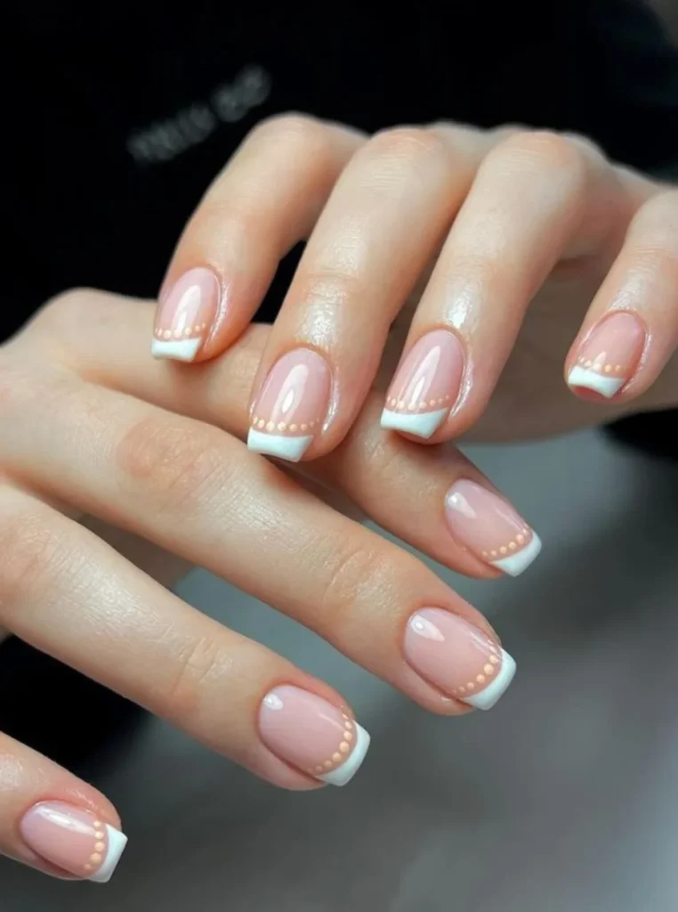 Elegante kurze French Nägel mit glänzender Nude-Rosa Basis, klassischen weißen Spitzen und feinen goldenen Punkten auf gepflegten femininen Händen.
