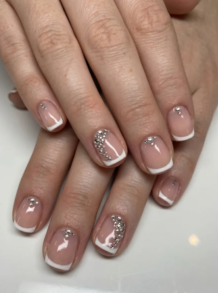 Elegante kurze French Nägel mit weißen Spitzen, Nude-Basis und funkelnden Strasssteinen in stilvoller Nahaufnahme.