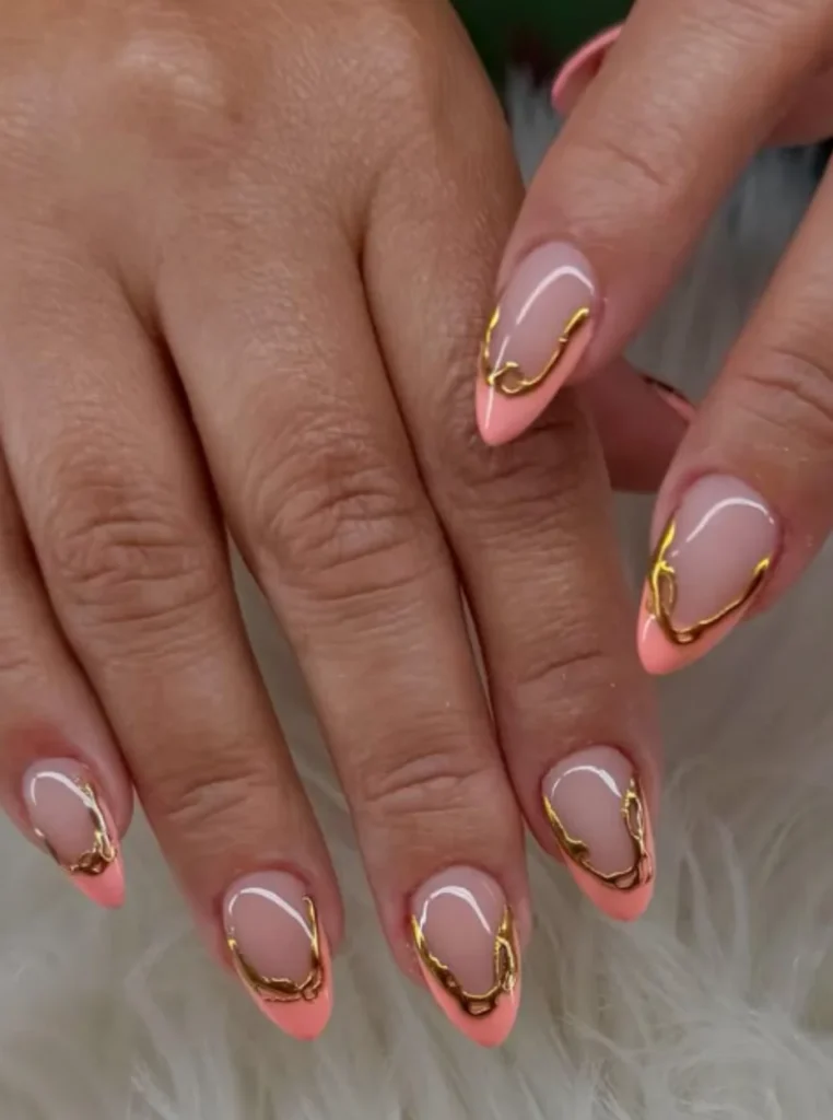 Apricot French Tips mit goldenen Akzenten auf mandelförmigen Nägeln und eleganter Nude-Basis.