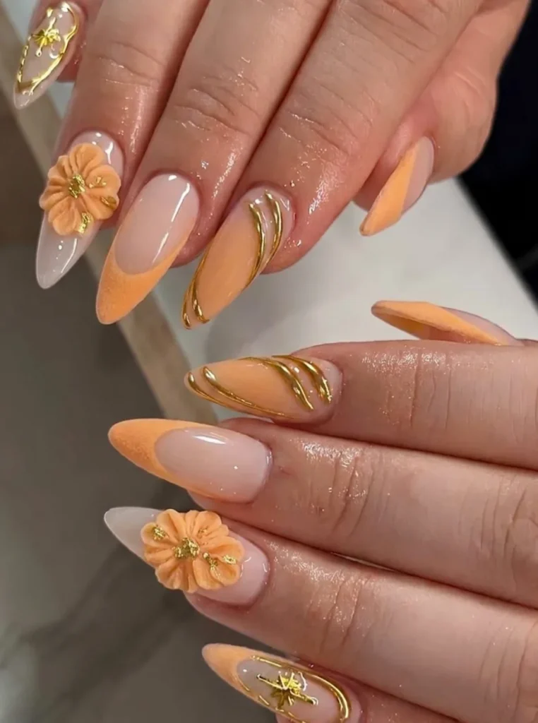 Apricot Nägel mit Goldfolie, 3D-Blumen und eleganten goldenen Verzierungen auf mandelförmigen Nägeln.