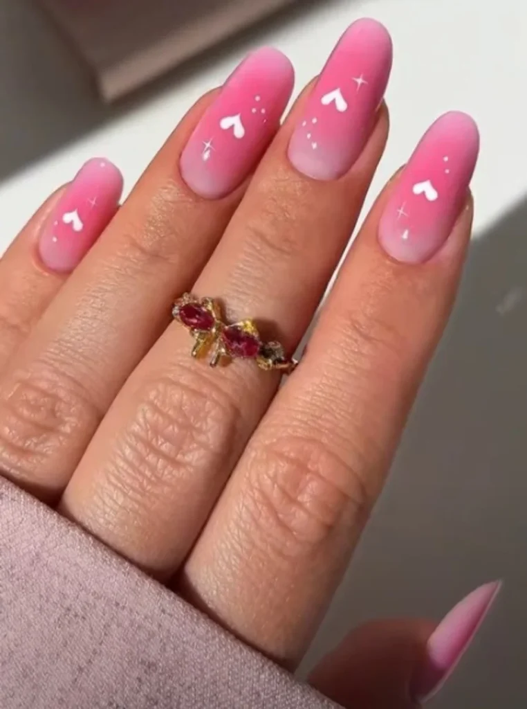 Aura Pink Nails mit sanftem rosa Farbverlauf, weißen Herzmotiven und mattem Finish auf mandelförmigen Nägeln.