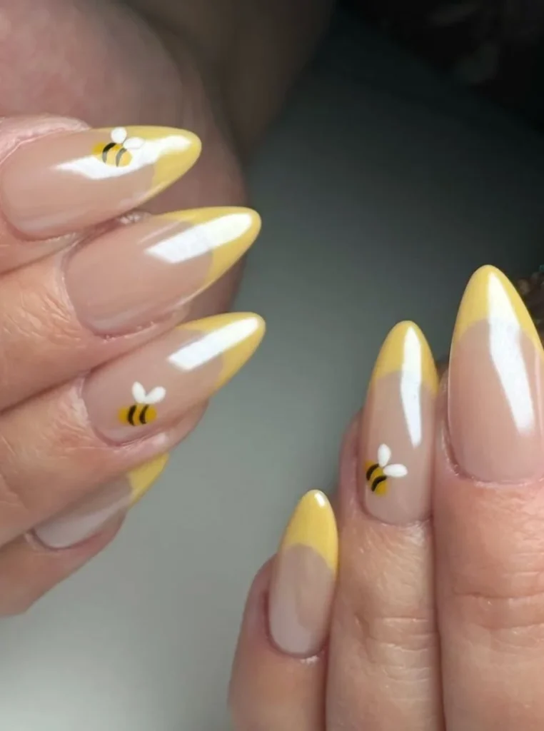 Chrome gelb French Nägel mit glänzenden gelben Spitzen und süßen Bienenmotiven auf mandelförmigen Nägeln mit Nude-Basis.