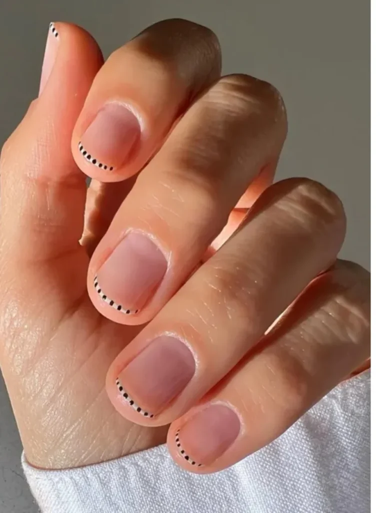 Minimalistische kurze French Nägel mit zarter Nude-Basis und feinen schwarzen Punktdetails entlang der Nagelspitzen für einen modernen und eleganten Look.