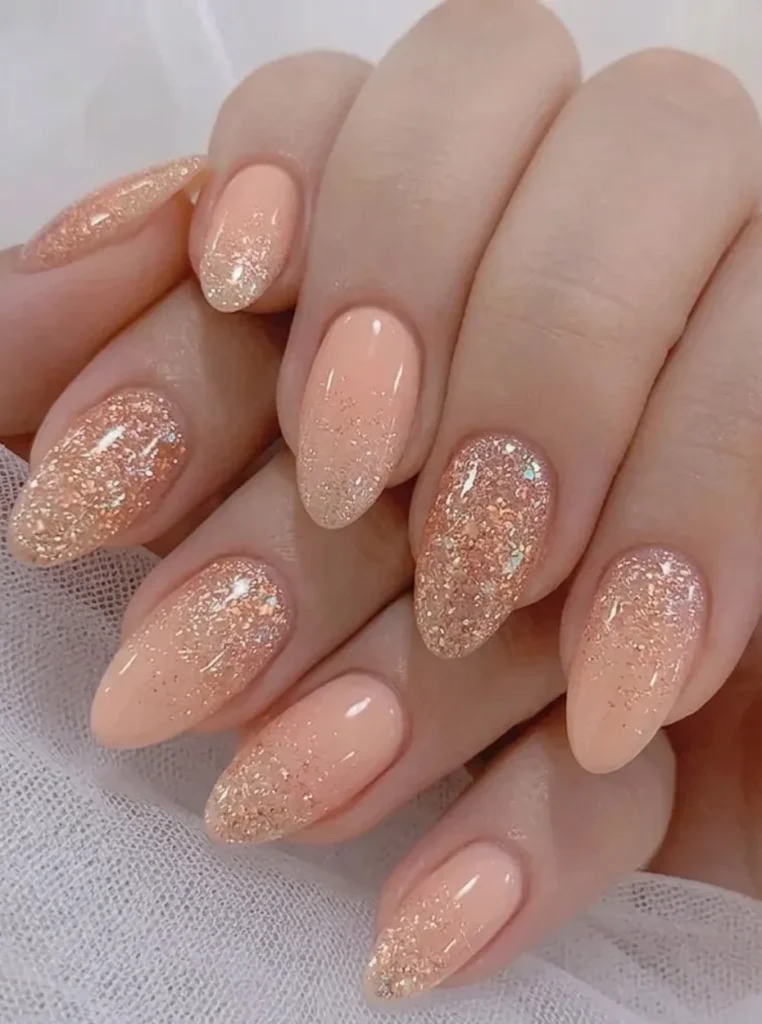 Pfirsich Nägel mit funkelndem Glitzerverlauf, schimmernden Akzenten und elegantem Ombre-Effekt auf mandelförmigen Nägeln.