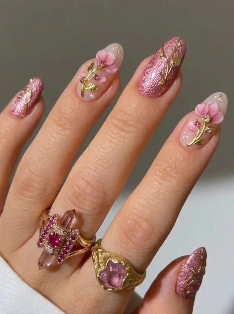 <Rosa 3D-Blumen Nails mit floralen Verzierungen, goldenen Details und strukturiertem Metallic-Finish auf mandelförmigen Nägeln.